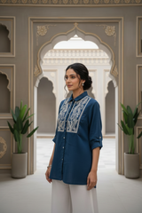 Woman in a blue embroidered shirt standing in an ornate architectural setting with plants.